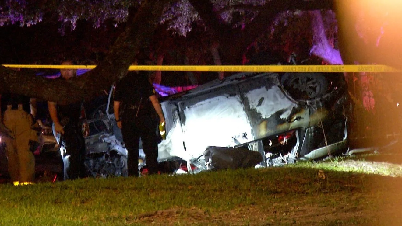 Police investigate a burned SUV crash scene near a fence and covered parking structures in San Antonio